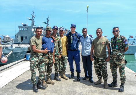 Chris Salinas is flanked by men in fatigues, and they stand in front of a ship in Timor-Leste