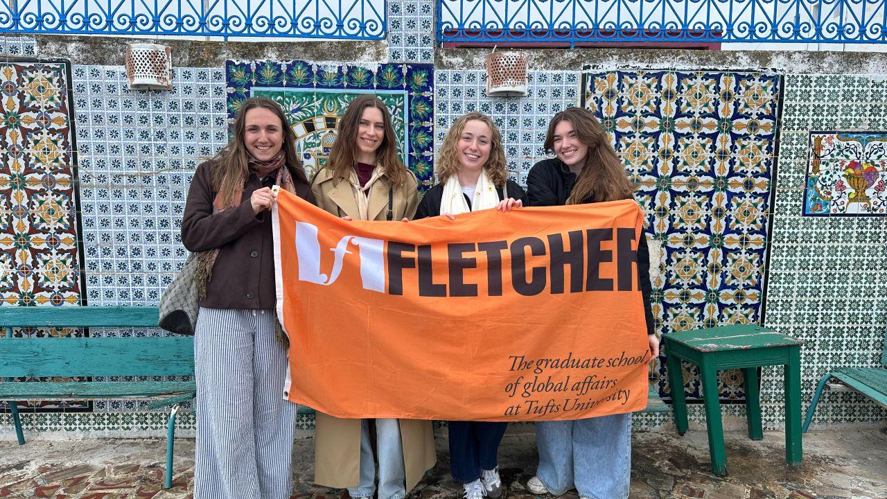 Four students hold the Fletcher flag in front of a tiled wall in Tunisia.