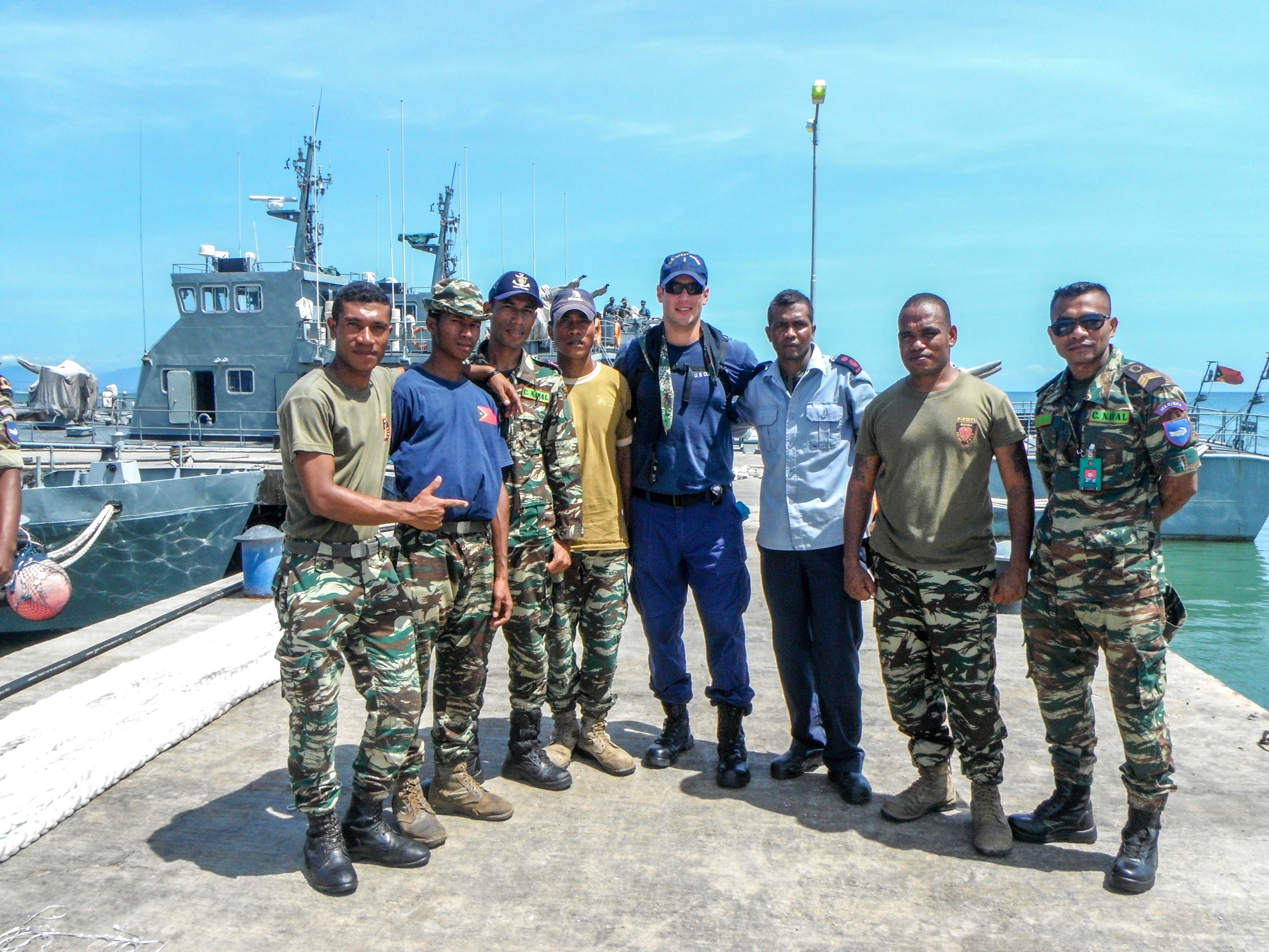 Chris Salinas is flanked by men in fatigues, and they stand in front of a ship in Timor-Leste