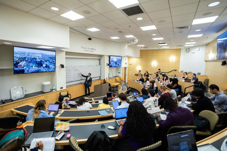 A wide angle picture of a classroom, students sitting in curved desks around a professor pointing at the white board