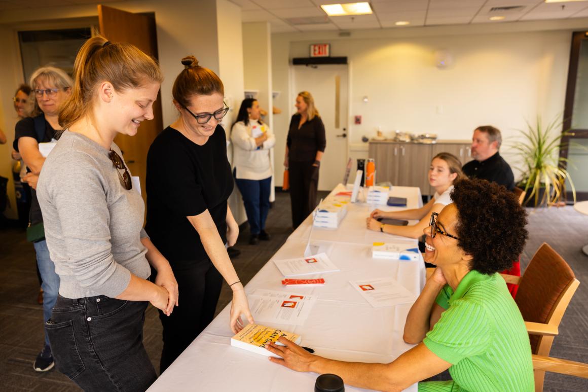 Two women stand and smile down at another woman sitting down. They are exchanging a book.