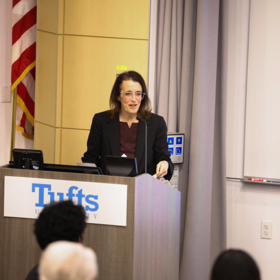 Kelly Sims Gallagher smiles while delivering remarks behind a Tufts podium.