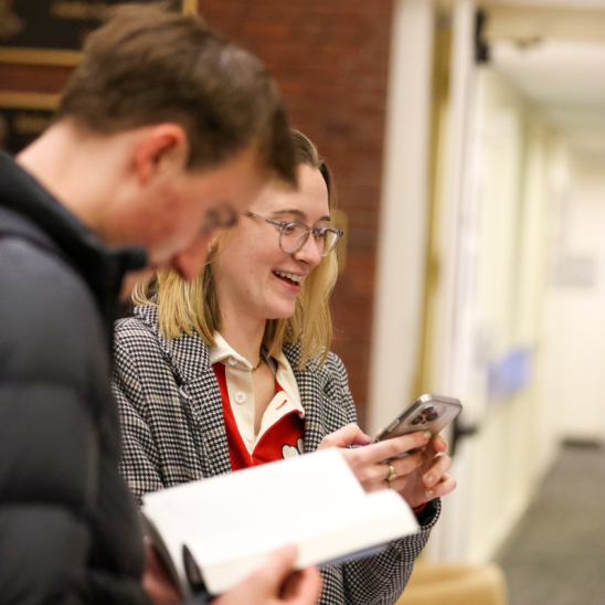 A student smiles while looking at her phone while another reads a book.