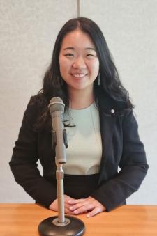 A headshot of Deborah Jeong standing at a podium before a microphone