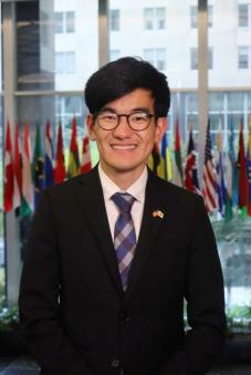 A headshot of John Cho in front of flags in the background