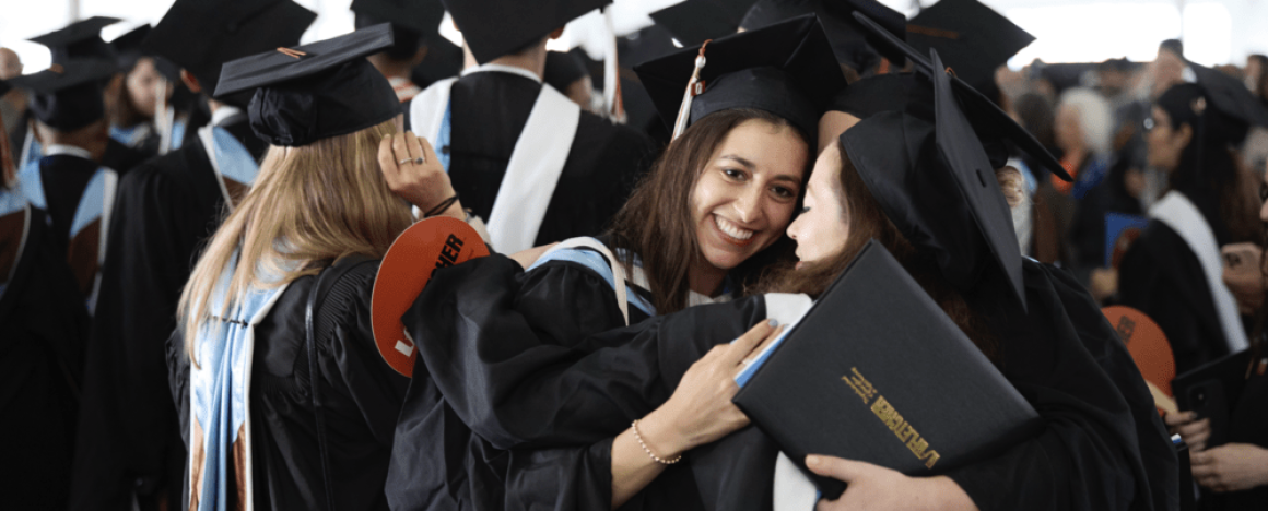 Two Fletcher graduates in black caps and gowns hugging in front of a crowd of many graduates
