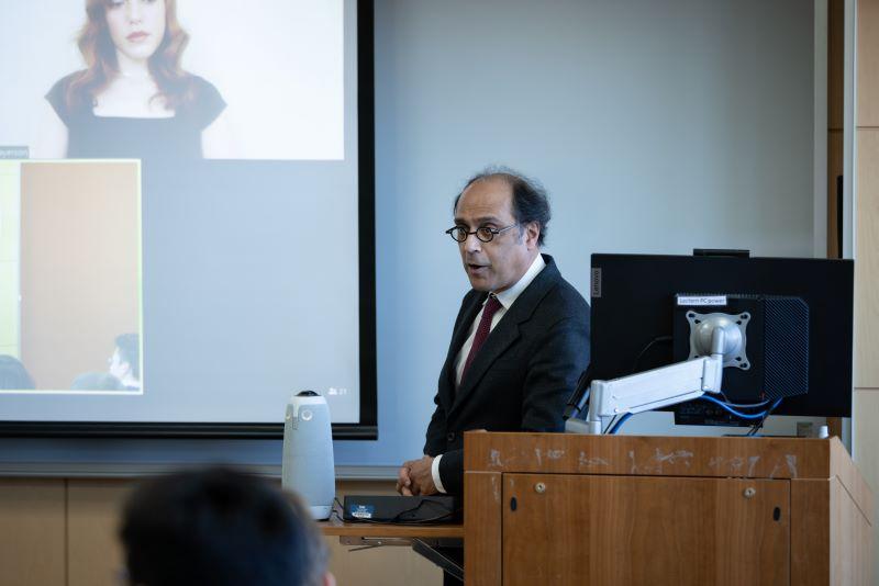 Dean Chakravorti speaks at the podium, and Zoom panelists can be seen on a screen in the background.