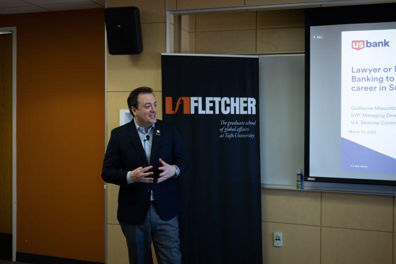 Guillaume Mascotto stands speaking in front of a Fletcher School banner and a screen