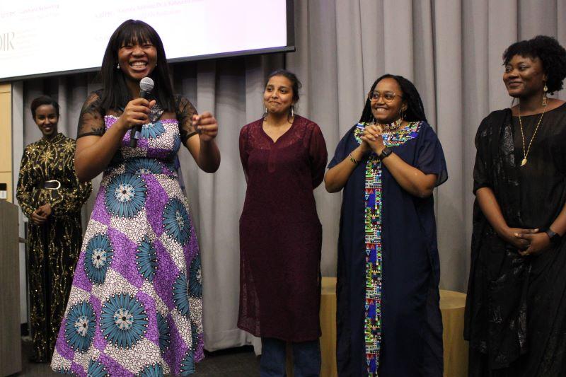 A group of students stands in the front of a room while one speaks.