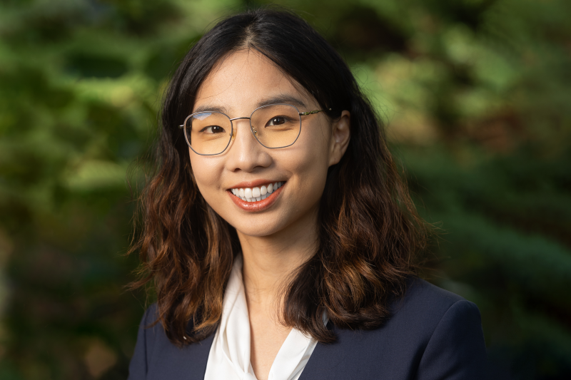 A headshot of Sally Zhang in front of a blurred, green, outdoor background.