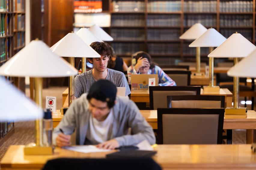 Students are working in a library. Two are on laptops and one is writing on paper.