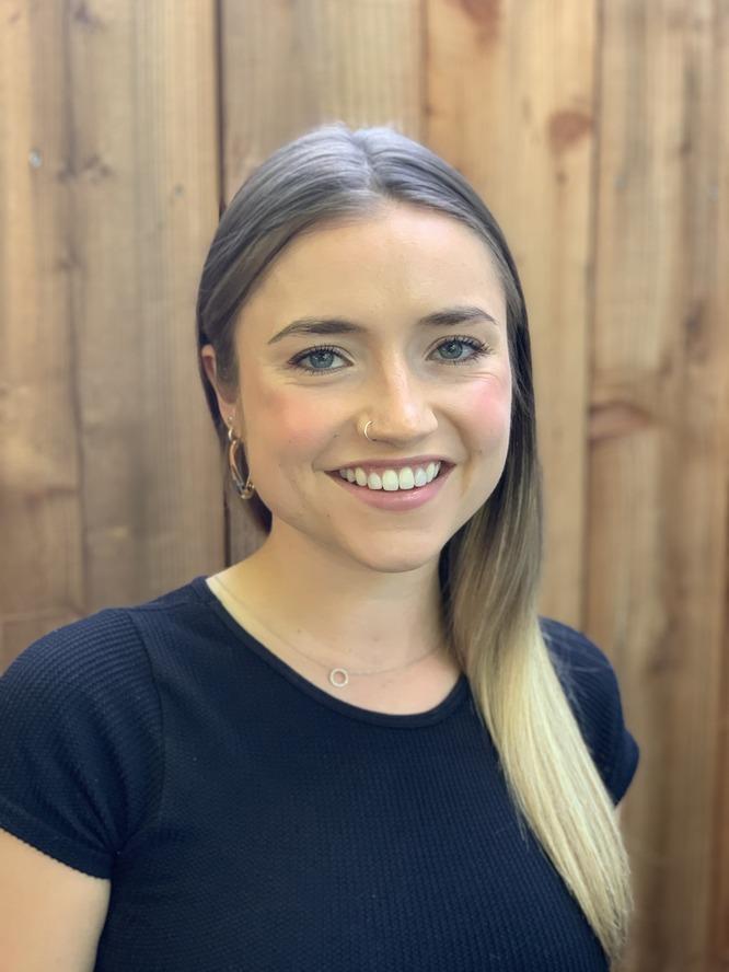 A person with blond hair and wearing a navy blue tshirt smiles in front of a wooden background.