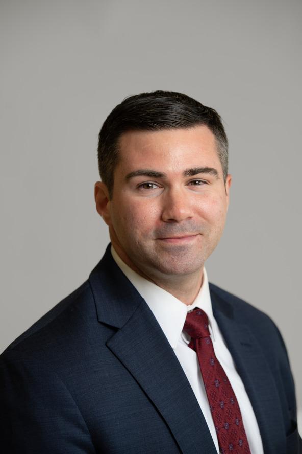 A person with short black hair smiles in front of a gray background wearing a suit with a red tie.