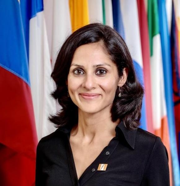 Avantika wears a v-neck black top with a Fletcher pin and smiles in front of assorted flags.
