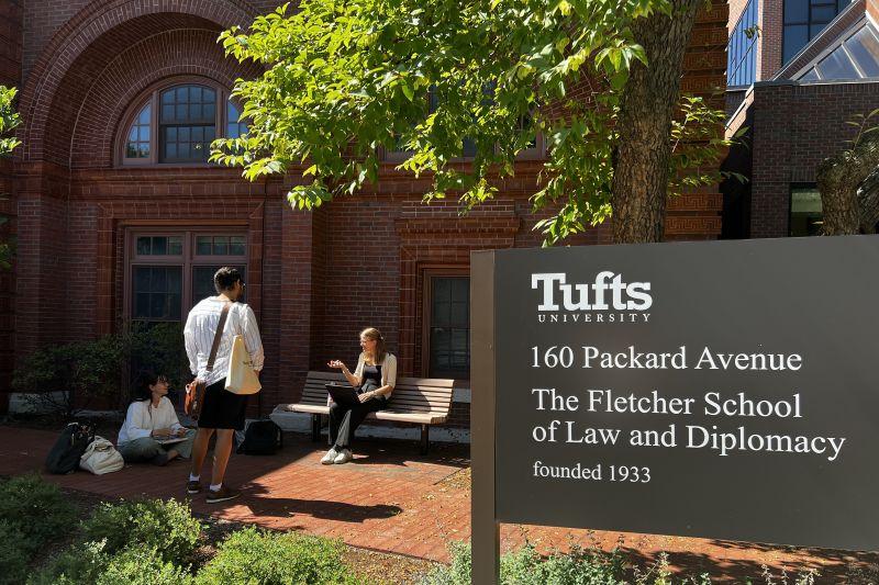 Three Fletcher students sit outside of a building on Fletcher's campus with a Tufts sign in the foreground.