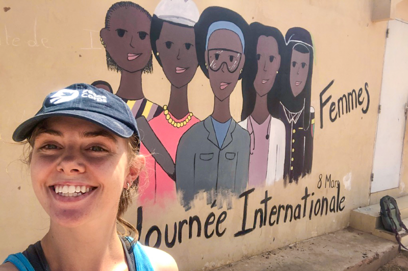 Emily Ruhm smiles in front of a mural of five women, which says, "Journee Internationale"