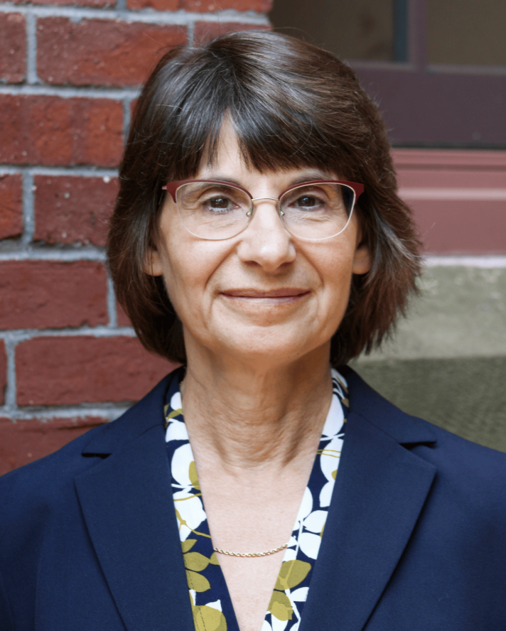 Janet wears glasses and a navy suit and smiles in front of a brick wall