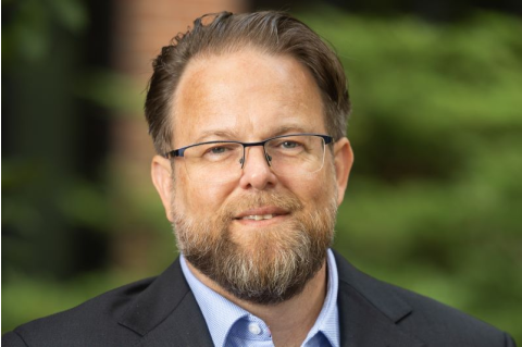 A headshot of Adam Kamradt-Scott, smiling in front of a blurred, green, outdoor background.