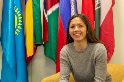 Eliana Johnson sits in a chair, smiling, in front of a background of flags. 