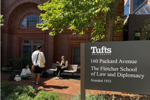 Three Fletcher students sit outside of a building on Fletcher's campus with a Tufts sign in the foreground.
