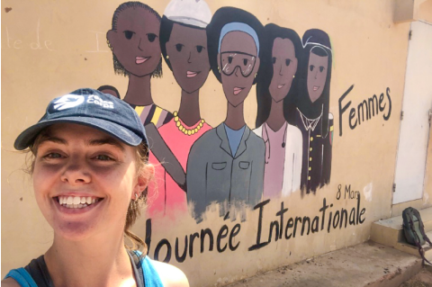 Emily Ruhm smiles in front of a mural of five women, which says, "Journee Internationale"