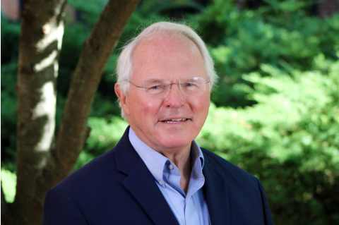 Ambassador Christopher Hill smiles for a headshot in front of a blurry green background outdoors. 