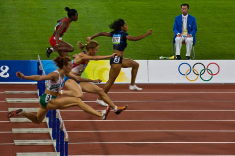 Women jumping hurdles in Olympic competition, with mad sitting on chair watching them. The richer the country, the more likely it will tally more Olympic medals—and the number of medals for women is tied to their economic opportunities at home
