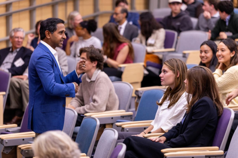 A man in a blue suit stands in front of a room of students, speaking with them.